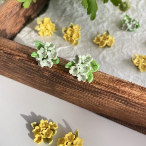 Mint white floral bouquet stud earrings with green leaves on a wooden tray, with yellow flower studs and daisies in the background, soft natural sunlight.